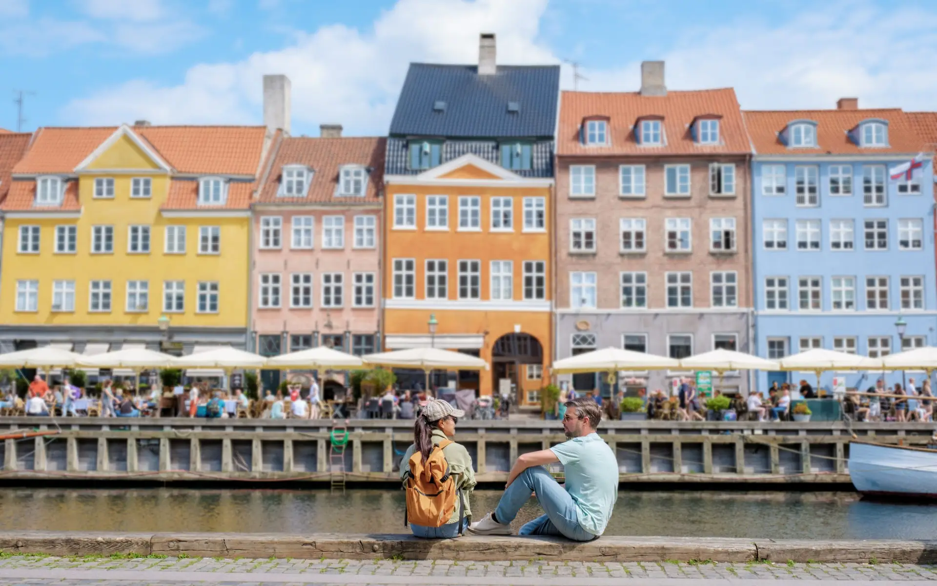 Where is Copenhagen located? Where is Copenhagen located? This charming scene captures a couple relaxing by the vibrant canal, surrounded by colorful buildings and a lively atmosphere.