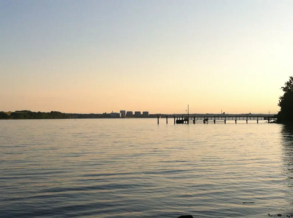 Kolding Fjord: Experience Denmark’s Living History Amidst Stunning Natural Beauty. A calm view of Kolding Fjord at sunset, with still waters, a wooden pier, and the city skyline in the distance.