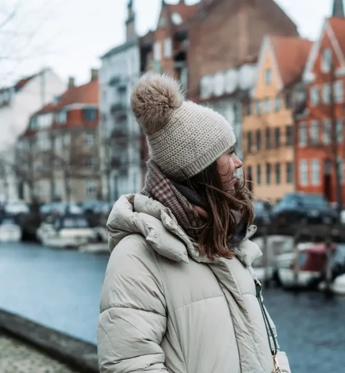 Does it snow in Copenhagen? A woman wearing a winter jacket and scarf, walking by the canal in Copenhagen, highlighting the city's cold weather during the winter season.